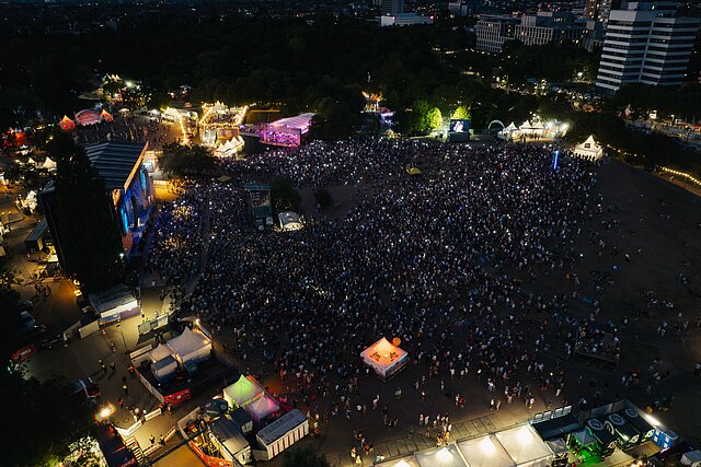 Weite Luftaufnahme des Festivalgeländes von DAS FEST Karlsruhe am Abend mit Bühnen, Zelten und tausenden Besuchern
