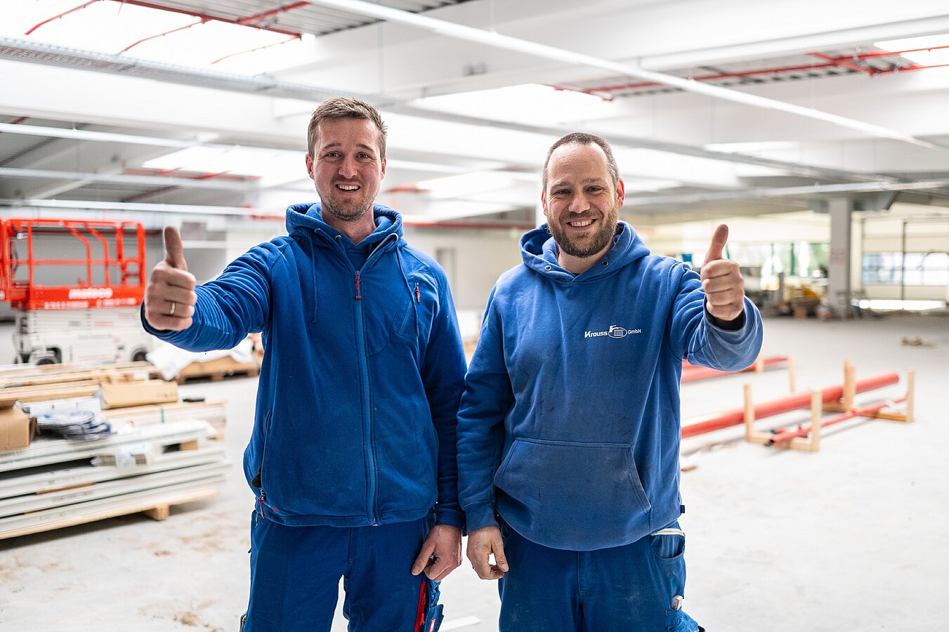 Zwei Monteure zeigen lachend Daumen hoch auf einer Baustelle