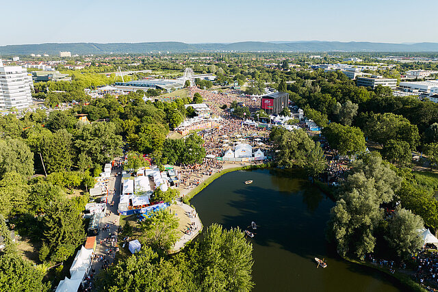 Luftaufnahme des Festivalgeländes von DAS FEST Karlsruhe im Abendlicht mit Bühne, Riesenrad und See