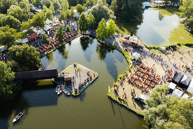 Drohnenaufnahme von oben auf den Festivalsee mit Insel, Holzbrücke und Ruderbooten bei DAS FEST Karlsruhe