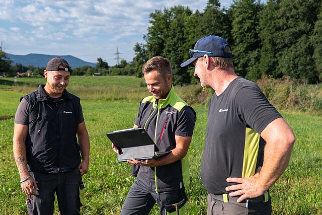 Drei Zaunteam-Monteure besprechen die Zaunplanung am Tablet, im Hintergrund die Berge im Chiemgau