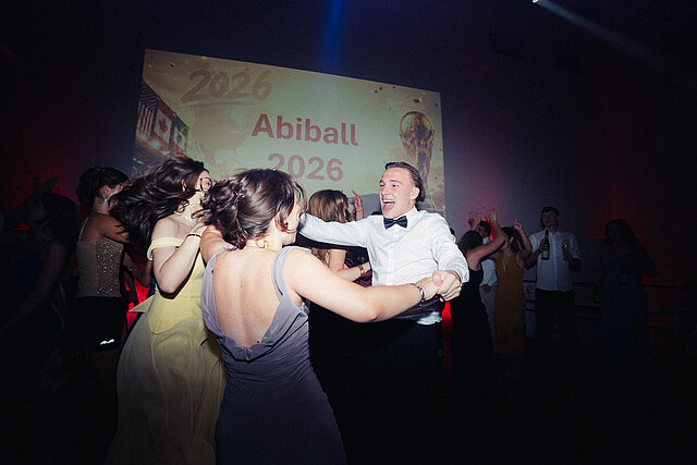 Abiturient mit Fliege tanzt mit zwei Freundinnen vor der Leinwand Abiball 2026 mit WM-Pokal